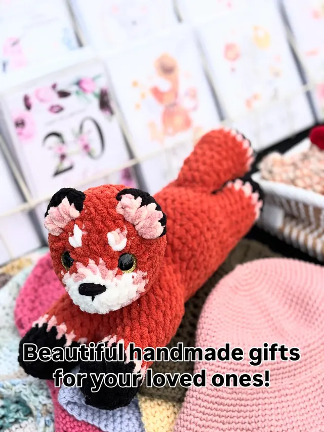A soft rust-red crochet fox lying on a pile of crochet pieces, with greeting cards behind it.