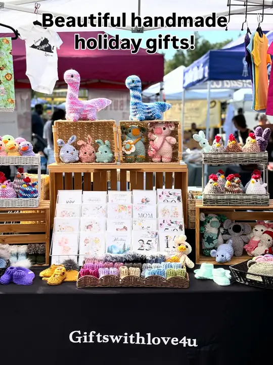 A Giftswithlove4u market table with crochet dinosaurs, bunnies, chickens and bears in baskets, rows of greeting cards, and baskets of crochet baby booties.