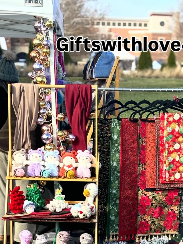 The Giftswithlove4u stall outdoors in winter, with shelves of crochet geese and animals beside a rack of holiday fabric pieces.